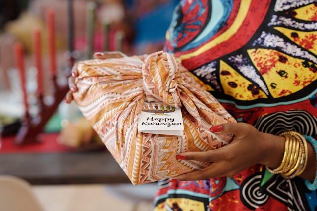 Femme qui tient un cadeau emballé dans un furoshiki avec une étiquette "happy kwanzaa"