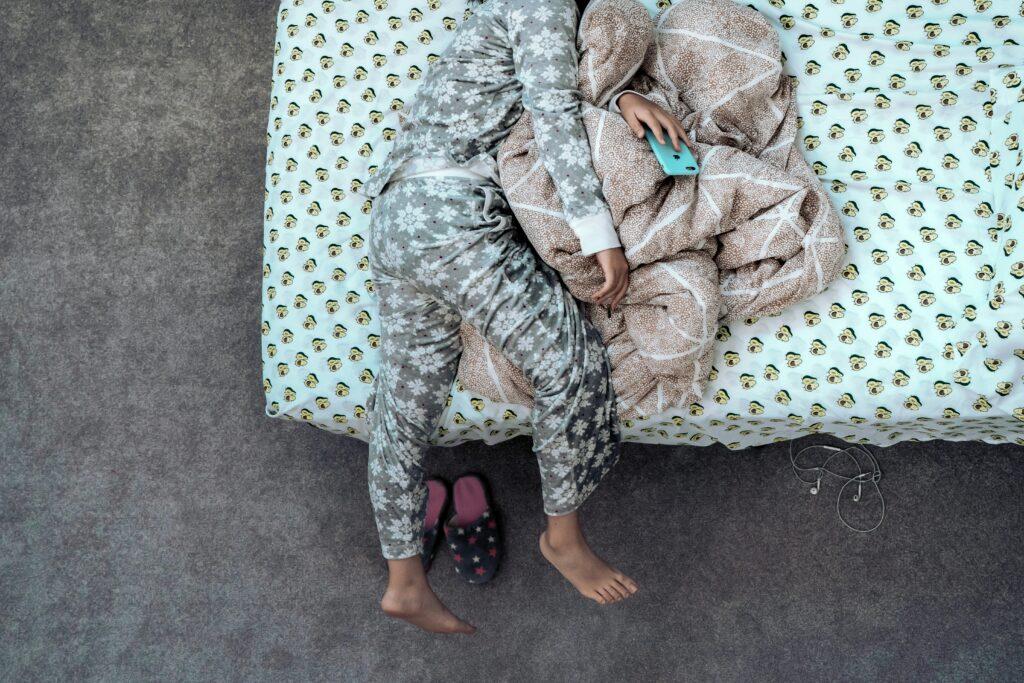 Photo d'une personne en pyjama affalée sur un lit avec un plaid et des pantoufles.