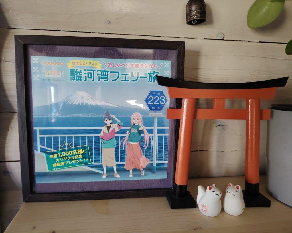L'image montre un cadre avec une affiche récupérée dans une gare au Japon, où on voit les personnages de l'anime Yuru Camp devant le mont Fuji. Le cadre est posé sur une étagère à côté d'une réplique de torii japonais et de deux figurines ramenées du Japon.